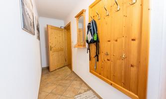 A bright hallway with tiled flooring and a wooden hook system on the wall. A backpack is hanging on the wall and the entrance door is visible.