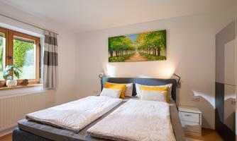 A cozy bedroom with two single beds and light colors. A picture of a tree-lined avenue hangs on the wall, and the window provides daylight.