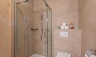 A modern shower cabin with glass doors in a bright bathroom. The walls are tiled in neutral tones.