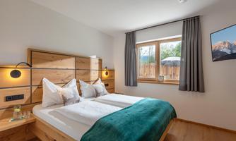 A cozy bedroom with a double bed and wooden details. There is a window overlooking nature and soft curtains.