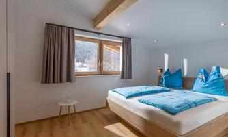 A modern bedroom with a large window overlooking the mountains. There is a double bed with blue pillows and a small table next to the bed.