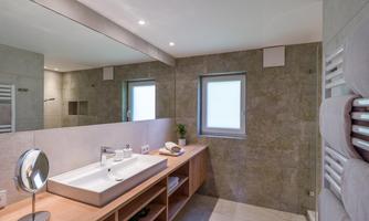 A modern bathroom with a large mirror and a sink made of white materials. It has a bright and simple decor with wooden shelves.