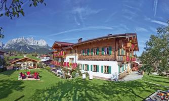 A charming wooden house with colorful flower balconies, surrounded by green lawn. In the background, majestic mountains and a clear sky can be seen.