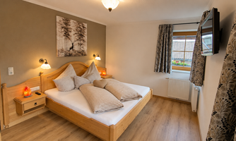 A cozy bedroom with a double bed and wooden furniture. The walls are painted in soft colors, and a window offers a view outside.