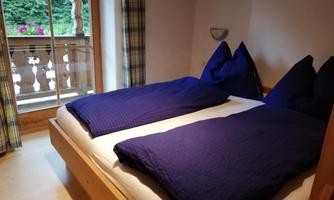 A cozy bedroom with two single beds and blue bedspreads. The room has a balcony with a view of greenery.