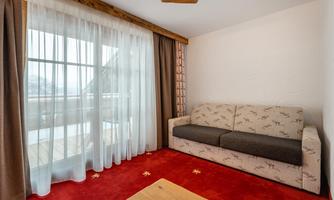 A cozy living room with a gray sofa and a red carpet. Large windows with curtains provide plenty of daylight.