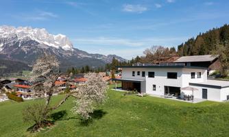 A modern house in a green landscape with a view of the mountains. The surroundings are picturesque with trees and small villages nearby.