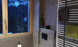 A modern bathroom with a white toilet and a large window pane. The walls are made of gray stone and there are towels on a radiator.