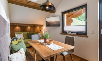 A cozy living space with a wooden table and green sofas. The large windows offer a beautiful view outside and the room is bright and inviting.