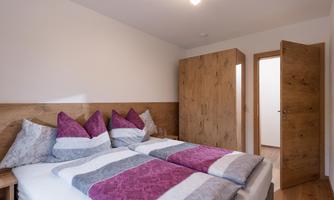 A cozy bedroom with a double bed and colorful pillows. The room has a warm wood look and an open door to an adjacent room.
