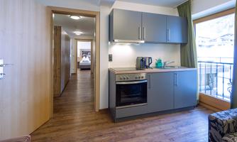 A modern kitchenette with a stove and coffee maker. In the background, a bright room with access to the balcony is visible.