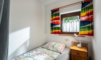 A small, bright room with a single bed and colorful curtains. On the nightstand, there is a lamp and the window lets in plenty of light.
