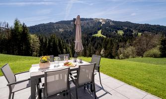 A beautiful outdoor area with a table and chairs on a meadow. In the background, there are wooded hills and a blue sky.