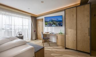 A modern hotel room with a large window and wooden details. The room features a bed, a desk, and a TV.