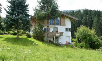 A modern house surrounded by green grass and trees. In the background, the mountains and a forest are visible.