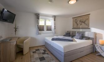 A modern bedroom with a large bed, a desk, and a television. There are large windows that let in plenty of natural light.
