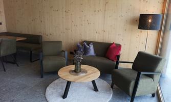 A cozy living area with a round table and comfortable armchairs. The walls are made of light wood, and there is a modern lamp in the background.