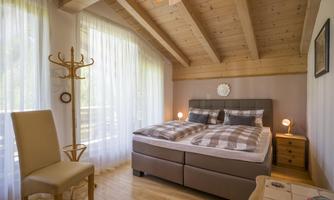 A cozy bedroom with wooden beams, a large bed, and soft colors. There is a window with curtains and a chair next to a clothes rack.