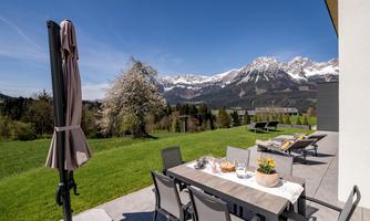 A cozy dining table on a terrace with a view of snow-covered mountains. The garden is green and a sun umbrella is ready.