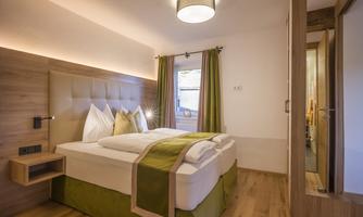 A modern bedroom with a large bed and elegant bedding. The walls are bright, and large windows provide pleasant lighting.