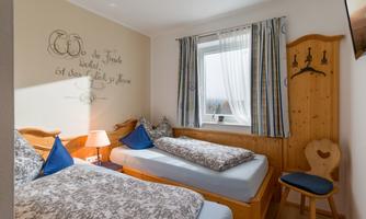 A cozy bedroom with two single beds, blue cushions, and a wooden chair. A window lets in plenty of light, and there is a decorative saying on the wall.