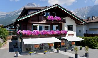 A charming, traditional house with colorful flowers on the balcony. In the background, mountains and a clear sky can be seen.