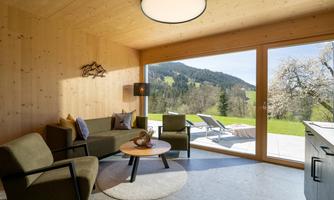 A modern living room with a large window front and a view of green hills. The decor is minimalist and cozy, with a round table and comfortable chairs.