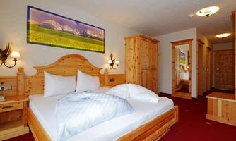 A cozy hotel room with wooden furniture and a large bed. On the wall hangs a picture of mountains and a green meadow.