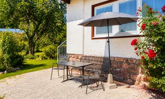 A cozy outdoor area with a table and chairs under a sunshade. Surrounded by green plants and flowers, it offers an inviting place to relax.