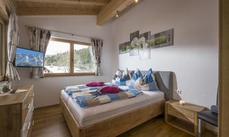 A modern bedroom with a large window and mountain view. The room is cozily furnished, with a double bed and a television.