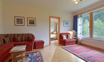 A cozy living room with a red sofa and wooden floor. Large windows offer a view of the outside, decorated with pictures on the walls.