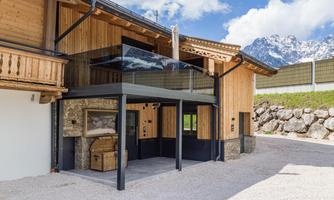 A modern chalet with wood and stone elements in a rural setting. In the background, mountains and a blue sky are visible.
