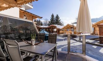 A cozy terrace with a table and chairs. In the background, snow-covered huts and trees can be seen.