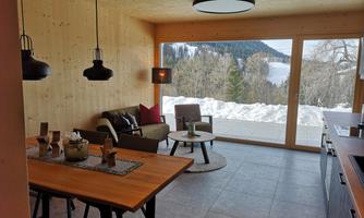 A modern living room with wooden walls and large windows that offer a view of snow-capped mountains. The room is cozily furnished with a dining table and a seating area.