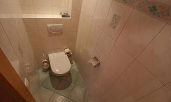 A small bathroom with a wall-mounted toilet and tiles. Basic hygiene items such as toilet paper and a towel holder are visible.