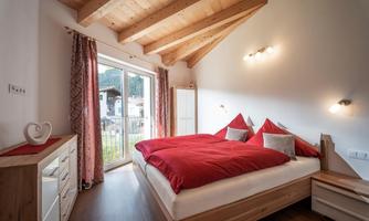 A cozy bedroom with a large bed and red pillows. Large windows let in a lot of light and offer a view outside.