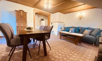 A cozy living room with a dining area and a comfortable sofa. The room has a warm wooden ceiling and inviting colors.
