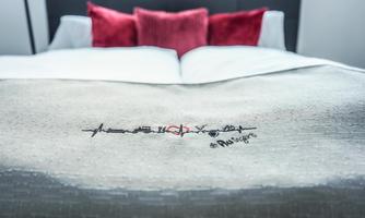 A bed with a white blanket and red pillows. On the blanket is a heartbeat pattern with the text "#RUgivers" embroidered.