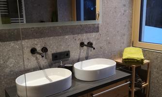 A modern bathroom with a double sink and two round basins. The walls are clad in grey stone, and there are large windows that let in natural light.