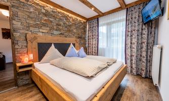 A cozy bedroom with a wooden bed and two pillows. The walls are clad with natural stone and a television is installed.