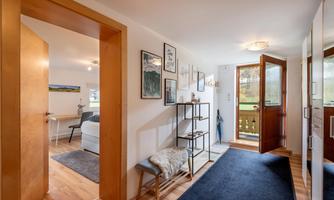 An inviting hallway with an open entrance and wooden-paneled walls. Pictures hang on the wall, and a shelf stands on the floor.
