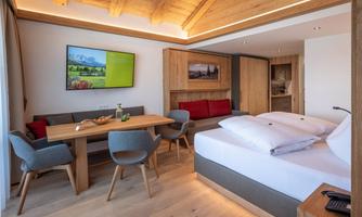 A modern room with wooden elements, a large bed, and a dining table. A television is mounted on the wall, and there are comfortable chairs for relaxation.