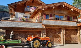 A rustic wooden house with blooming balcony plants. In front of it stands a bright orange tractor.