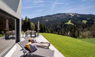 A modern terrace with loungers and colorful cushions offers a view of green hills and forests. The sky is clear and blue, perfect for relaxing days in nature.