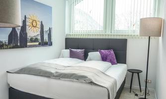 A modern bedroom with a cozy double bed and decorative pillows. A picture of monumental stones hangs on the wall, and large windows let in plenty of light.