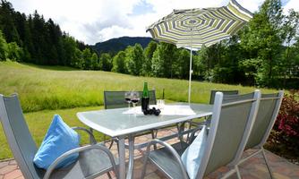 A beautiful table with chairs on a terrace, surrounded by green meadows and trees. A sun umbrella provides shade, and there are glasses and a bottle of wine ready.