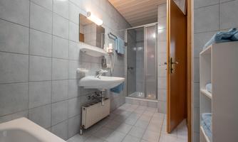 A modern bathroom with gray tiles, a sink, and a shower. On the right side, there is a wooden door and a shelf with towels.