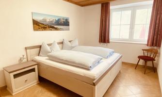 A cozy bedroom with a large double bed and a wooden floor. A picture of mountains hangs on the wall, and there is a chair next to the bed.