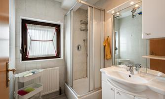 A modern bathroom with a shower and a sink. The walls are light tiled, and there is a window with a white curtain.