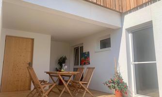 A cozy terrace with a wooden table and chairs. In the background, there are plants and a bright wall.